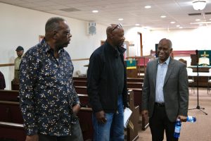 three men stand and talk. man on left is wearing dark shirt with prints, man in center is wearing dark jacket and jeans and man on right is wearing patterned sports coat