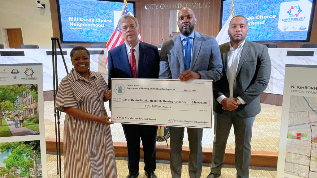 Four people stand holding a large cardboard check. There is a woman at far left and three men, all wearing suits. The check is for $50 million from the U.S. Department of Housing and Urban Development.