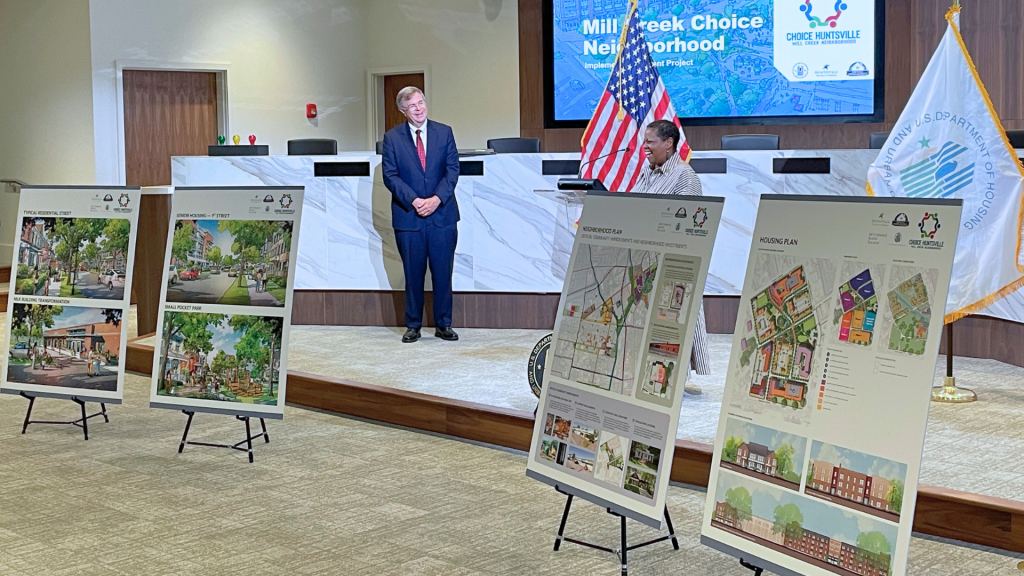 A woman speaks at a podium while Mayor Tommy Battle looks on. There are two flags behind her and four posters in the foreground showing renderings of the redeveloped Mill Creek area.