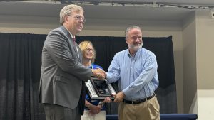 Huntsville Mayor Tommy Battle presents a plaque at the 49th annual Huntsville Beautification Awards on Aug. 22. He is at left in a gray suit and is shaking hands with a man in a blue shirt and brown pants. There is a woman standing in the back in a blue shirt.