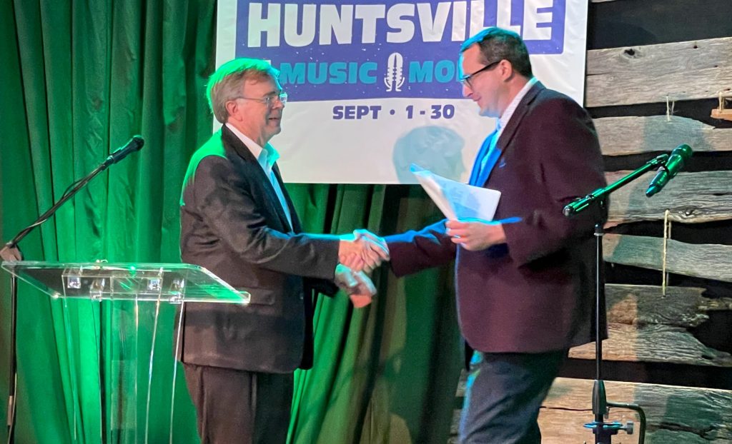 Mayor Tommy Battle welcomes Music Commissioner Matt Mandrella to speak at the Huntsville Music Month 2024 kickoff event at Tangled String Studios. Both men are wearing blazers. A large sign that reads 