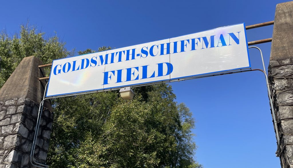 white sign with blue letters that says Goldsmith-Schiffman Field with large green tree and blue sky in background