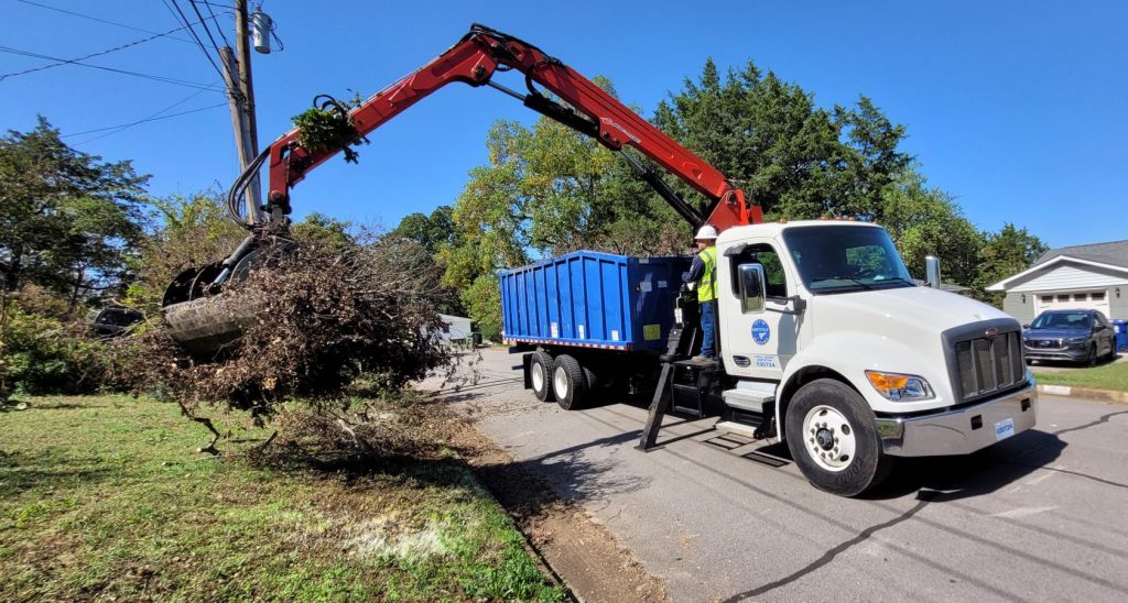 large truck with red arm extending to ground to pick up yard debris with green trees and blue sky in background