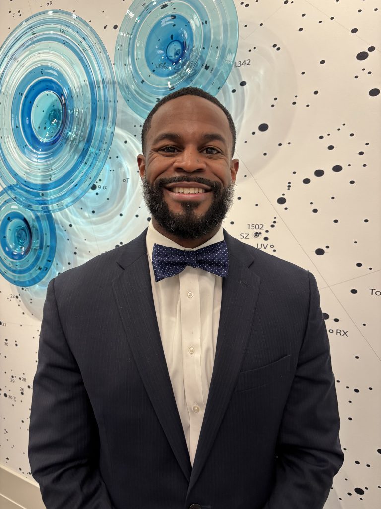 Smiling man wearing a suit and bowtie stands in front of glass artwork hanging on a wall