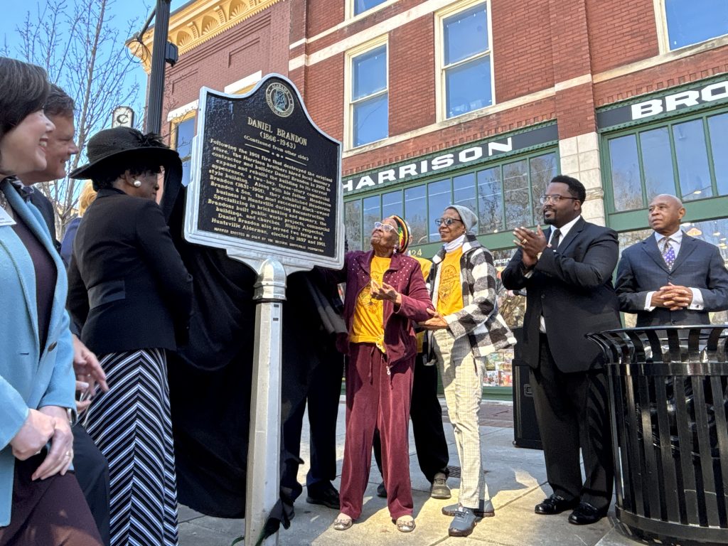 The dignitaries surround a historic marker after its unveiling. The marker, ahead of Harrison Brothers Hardware, in downtown Huntsville, presents the story of Henderson and Daniel Brandon. People in the photo look at the marker and applaud.